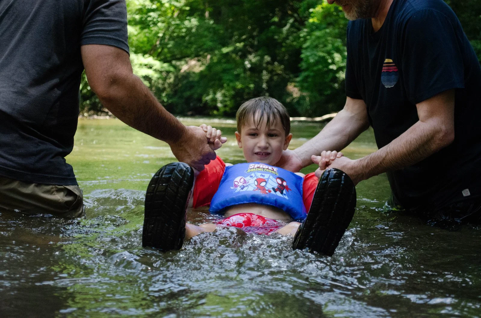 A smiling boy in a life vest floats in the river, held by two leaders from Ellijay City Church