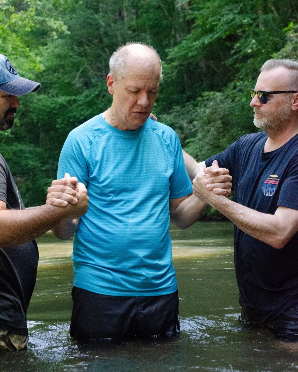Two church leaders hold a man by the hands as he prays, standing waist-deep in the river for his water baptism